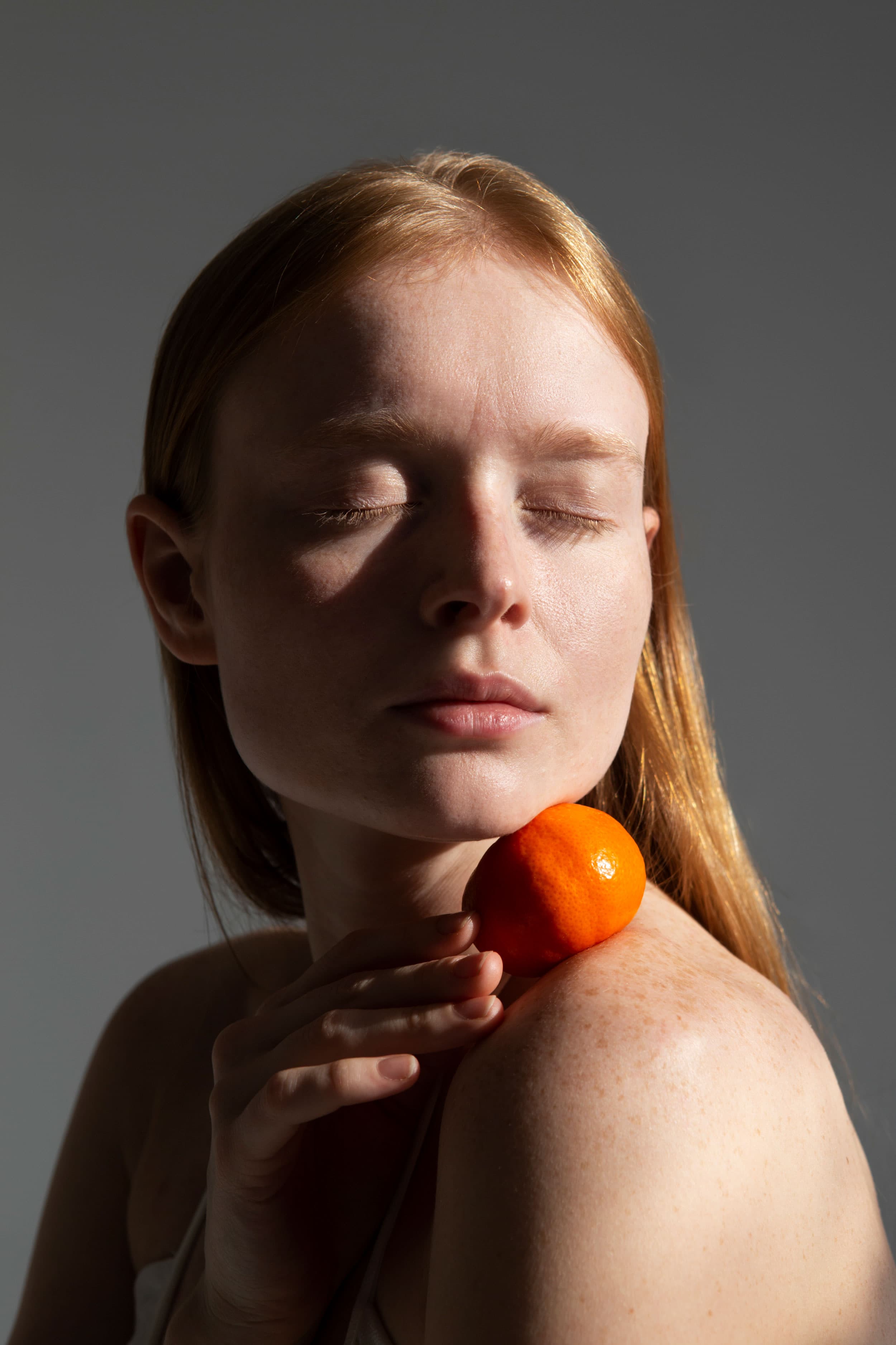 Before — redhead model holding a tangerine, unretouched, with subtle redness and visible imperfections